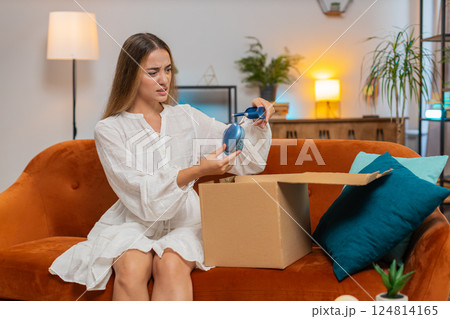 Dissatisfied annoyed young woman opening cardboard box receive broken wireless headphones at home 124814165