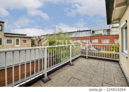 A spacious modern balcony featuring a railing, overlooking an urban environment with buildings and greenery, under a cloudy sky. 124814264