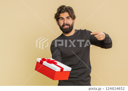Smiling young Indian man presenting birthday gift box stretches out hands against beige background 124814652