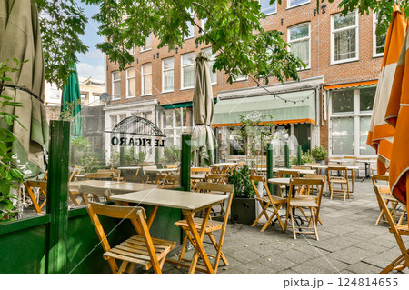A quaint outdoor cafe scene features wooden tables and chairs nestled among lush greenery, with colorful umbrellas adding a vibrant touch to the setting. 124814655