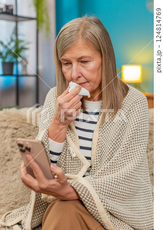 Mature woman covered with blanket on sofa, ill and sick, using smartphone to order medicine at home 124814769