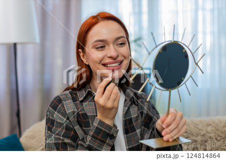 Young pretty woman applying makeup looking in mirror smiling while drawing lips with lipstick gently 124814864