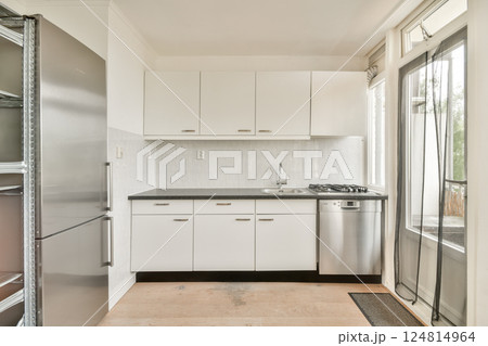 A minimalist kitchen featuring white cabinetry, stainless steel appliances, and a sleek countertop. Bright sunlight illuminates the space through large windows. 124814964