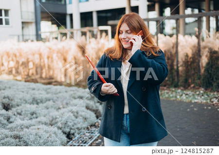 Young adult smiling professional business woman holding smartphone and folder with files while walking in urban city street. 124815287