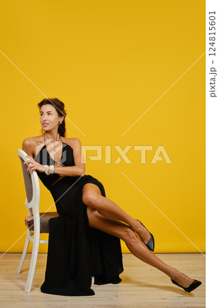Elegant woman in a black high-slit dress poses gracefully against a bright yellow backdrop Elegant woman in a black high-slit dress poses gracefully against a bright yellow backdrop 124815601