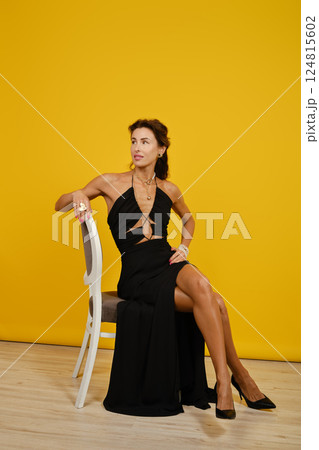 Elegant woman in black dress poses confidently against bright yellow backdrop Elegant woman in black dress poses confidently against bright yellow backdrop 124815602