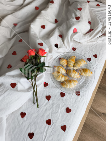 Romantic breakfast with golden croissants on crystal plate, red roses, and heart decorations on white bedding Romantic breakfast with golden croissants on crystal plate, red roses, and heart decorations on white bedding 124816043