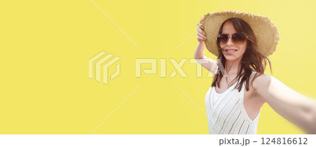 A smiling woman of 30-35 years old in a wide-brimmed hat and sunglasses on a yellow background, she takes a selfie. A concept for travel and blogging, copy space left background 124816612