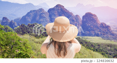 Woman female traveler in a wide hat shoots a rainforest landscape on a sunny day, rear view from the back. Panorama of the mountains of Khao Sok National Park Thailand, view from the observation deck Woman female traveler in a wide hat shoots a rainforest landscape on a sunny day, rear view from the back. Panorama of the mountains of Khao Sok National Park Thailand, view from the observation deck 124816632