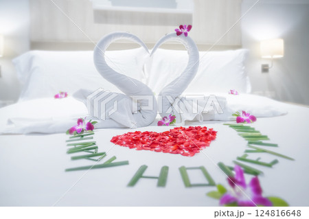 Swans made of hand-rolled towels, a symbol of love and relationships, and a heart made of rose petals on a white bed for lovers in a hotel room close-up. The Happy Honeymoon inscription is lined with 124816648