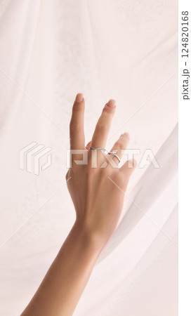 Cropped image of tender female hand with manicured nails wearing two delicate rings against a softly draped white fabric 124820168