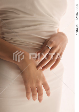 Hands of woman in elegant cream dress with solitaire engagement ring against neutral background. 124820200
