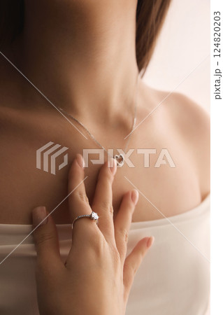 Close-up of woman hand with diamond engagement ring gently holding heart pendant on neck. 124820203