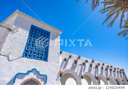 Nerja town, a resort of The Costa del Sol seaside region in Andalusia in the south of Spain. White building with blue details, flower pots, and arches against a bright blue sky. 124820494