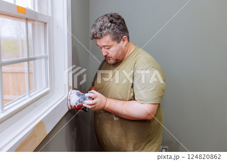 Wood worker carefully sands window trim preparing for further painting finishing in cozy interior space. Wood worker carefully sands window trim preparing for further painting finishing in cozy interior space. 124820862
