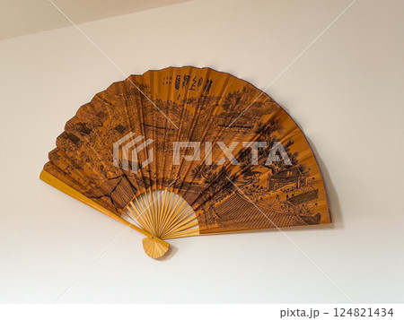 Traditional decorative fan on white wall with detailed historical landscape. Cultural heritage and visual storytelling through Asian artistry. Traditional decorative fan on white wall with detailed historical landscape. Cultural heritage and visual storytelling through Asian artistry. 124821434