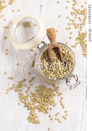 Glass jar full of raw green lentils with a wooden scoop on white table top view. Dried legumes 124824242