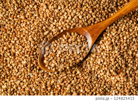 A wooden spoon filled with buckwheat grains rests on a surface covered with loose buckwheat seeds. The rich, earthy colors showcase the texture and natural elements of the grains 124825415