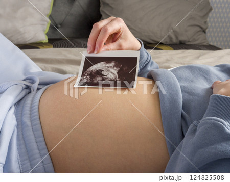 Pregnant woman resting on bed, cradling ultrasound image against growing belly, experiencing serene anticipation of upcoming maternal milestone 124825508