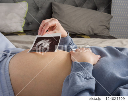 Expectant mother resting peacefully, holding ultrasound image against pregnant belly, experiencing emotional connection with growing baby during prenatal moment Expectant mother resting peacefully, holding ultrasound image against pregnant belly, experiencing emotional connection with growing baby during prenatal moment 124825509