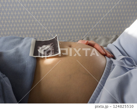 Pregnant woman relaxing, holding sonogram against growing belly, embodying maternal warmth and anticipation of new life Pregnant woman relaxing, holding sonogram against growing belly, embodying maternal warmth and anticipation of new life 124825510