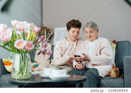 A young woman explaining her senior mother or grandma how to use smartphone. Retired lady discovering mobile technology for calling, texting, buying online, playing games, using medical apps. A young woman explaining her senior mother or grandma how to use smartphone. Retired lady discovering mobile technology for calling, texting, buying online, playing games, using medical apps. 124826954