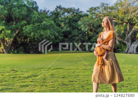 Mother holding her little baby in a yellow sling in the park. Warm and loving family moment. Babywearing, parenting, and mother-child bonding concept 124827880