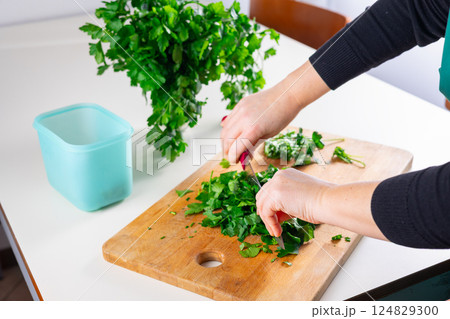 Close-up of a person hands chopping fresh parsley on a wooden cutting board in a kitchen.food container is on the table, ready to store the chopped herbs.Healthy food preparation and homemade cooking. Close-up of a person hands chopping fresh parsley on a wooden cutting board in a kitchen.food container is on the table, ready to store the chopped herbs.Healthy food preparation and homemade cooking. 124829300