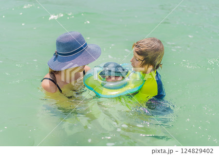 Baby floating in a neck ring in turquoise water with mom and older brother nearby. Family bonding and early swimming concept 124829402