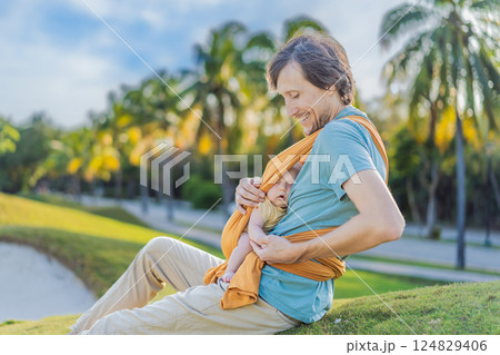Father holding his little baby in a yellow sling in the park. Warm and loving family moment. Babywearing, fatherhood, and parent-child bonding concept 124829406