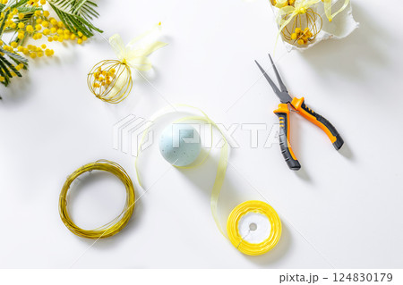 Top view of Easter craft materials on a white table: gold wire, yellow ribbon, pliers, mimosa flowers, and a decorative egg. Top view of Easter craft materials on a white table: gold wire, yellow ribbon, pliers, mimosa flowers, and a decorative egg. 124830179