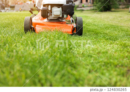 Gardening work tools. Wide angle view of orange electric lawn mower, wheels, motor on bright lush green grass at backyard. Rotary lawn mower machine cut lawn. Professional lawn care service. Gardening work tools. Wide angle view of orange electric lawn mower, wheels, motor on bright lush green grass at backyard. Rotary lawn mower machine cut lawn. Professional lawn care service. 124830265