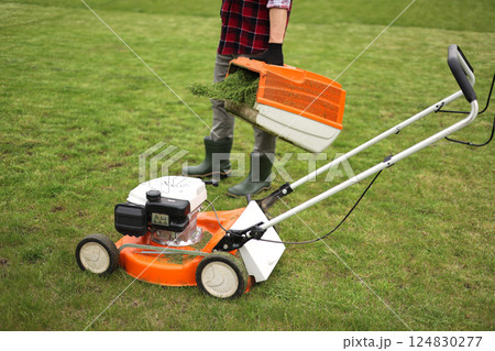 Unrecognisable man in casual clothes mows lawn with lawn mower at own backyard, holds bag with cut grass. Husband takes care of garden. Modern gasoline garden equipment. Landscaping work at home 124830277
