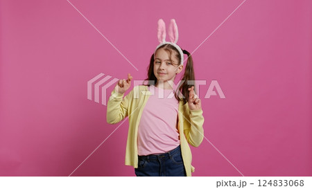 Lovely sweet girl posing in studio with fingers crossed and hoping to receive gifts for easter holiday celebration. Cheerful young child with bunny ears asking for good luck and fortune. Camera B. 124833068