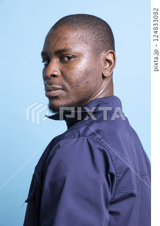 Portrait of african american security officer poses against blue background, looking over his shoulder in studio. Serious guarding personnel dressed in uniform, patrol duty. Portrait of african american security officer poses against blue background, looking over his shoulder in studio. Serious guarding personnel dressed in uniform, patrol duty. 124833082