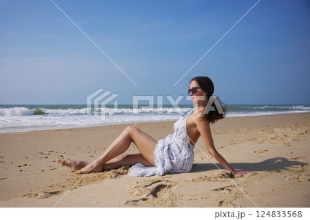 Girl in white summer dress sitting on beach near sea on a peaceful sunny day 124833568