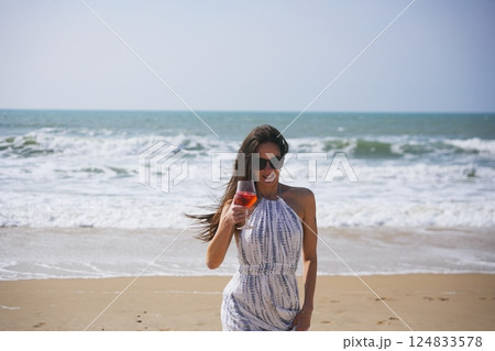 Stylish woman in summer dress holding cocktail on beach with ocean waves behind Stylish woman in summer dress holding cocktail on beach with ocean waves behind 124833578