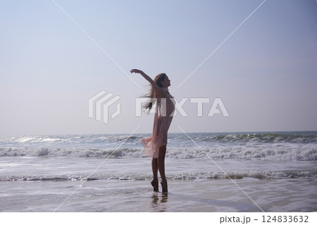 Ballerina on pointe at the ocean edge with arms raised and flowing dress 124833632
