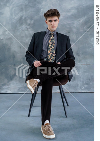 Young man in a dark suit, striped shirt, patterned tie, black pants, and beige sneakers casually seated on chair against gray textured wall 124840139