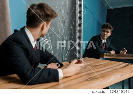Man in suit sitting at wooden table holding a smoking pipe while looking at his reflection in mirror 124840143