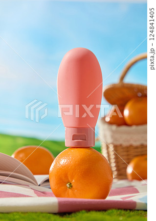 Pink elliptical tube of cream stacked on a round brilliant orange, with a checkered carpet underneath. The bamboo basket containing citrus burst orange is blurred in behind. Blue sky background. 124841405