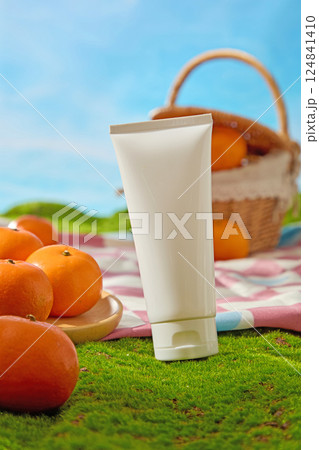 Close up of an unlabeled cream tube placed on green grass in the foreground. Round wooden plate containing fresh oranges with shiny peels. Natural light shining on objects in the garden.  124841410