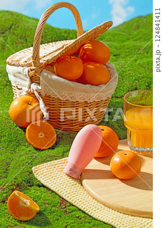 Vertical photo of a wicker basket with handles filled with fresh oranges. Unlabeled pink tube of cream sits next to a wooden cutting board and a glass of orange juice, displayed on a beige cloth rug 124841411