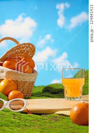 Glass of orange juice and white-rimmed sunglasses placed on a wooden cutting board, laid out on a dark green lawn. Fresh oranges with rough skins in a wicker basket. Blue sky with white clouds. Glass of orange juice and white-rimmed sunglasses placed on a wooden cutting board, laid out on a dark green lawn. Fresh oranges with rough skins in a wicker basket. Blue sky with white clouds. 124841413
