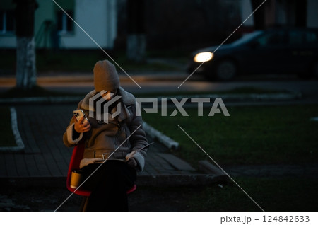 Woman seated on red chair in evening light, engaged with smartphone while reading notepad, surrounded by peaceful urban setting and passing car 124842633