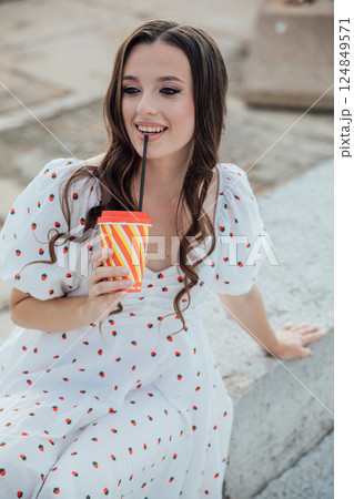 Beautiful slim fashionable brunette woman in summer dress sitting with a straw drink Beautiful slim fashionable brunette woman in summer dress sitting with a straw drink 124849571
