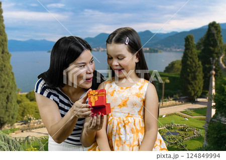 Surprised mother and her littler girl is opening a red gift box. Summer resort landscape background. Surprised mother and her littler girl is opening a red gift box. Summer resort landscape background. 124849994