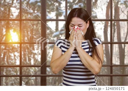 Asian woman with runny nose standing indoors. Sneezing brunette on checkered windows background with winter evening landscape. Asian woman with runny nose standing indoors. Sneezing brunette on checkered windows background with winter evening landscape. 124850171