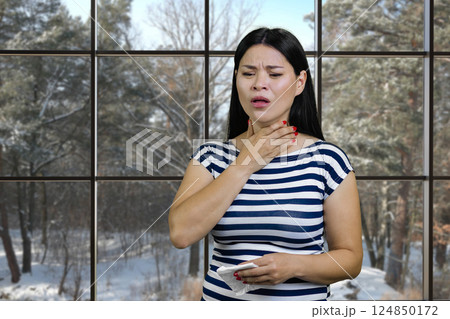 Young asian woman is suffering from cold touching her neck and holding a napkin. Ckeckered windows backgroud with view on a winter landscape. Young asian woman is suffering from cold touching her neck and holding a napkin. Ckeckered windows backgroud with view on a winter landscape. 124850172
