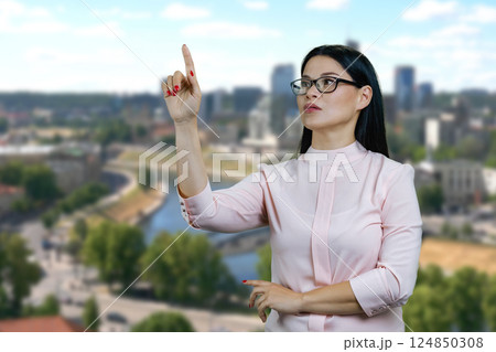 Young asian businesswoman is interacting with invisible virtual screen. Pointing at copy space. Blurred urban scape background. 124850308